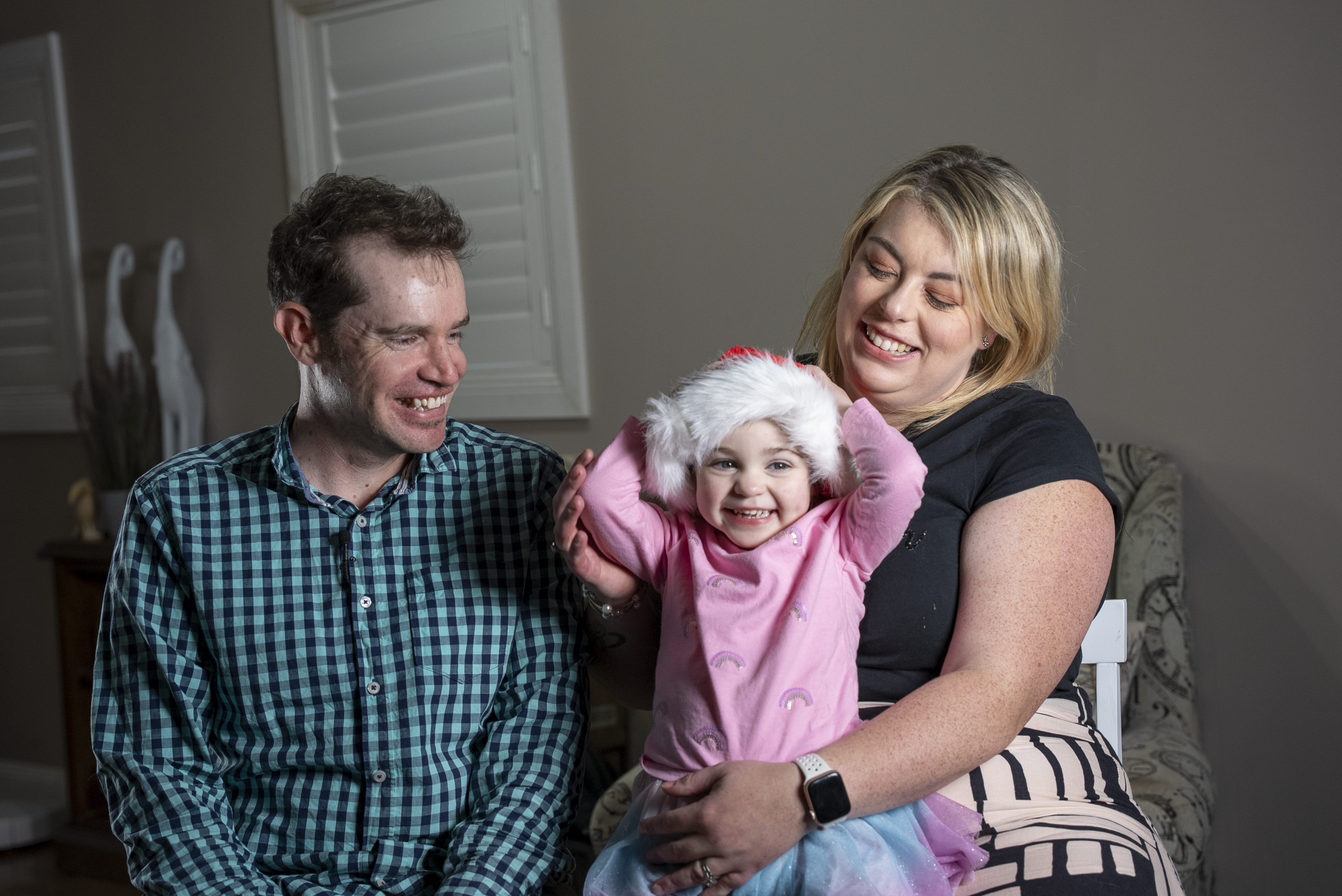 Image of child sitting on mother's knee, beside her father. Child wearing Christmas hat.