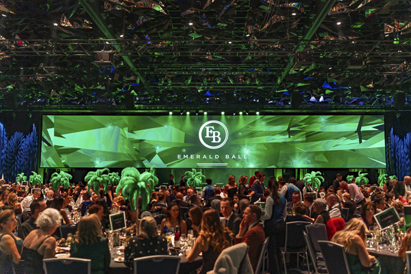 A view of the entire room at Emerald Ball 2025. Guests are sitting down at tables in front of a massive wide screen image of the Emerald Ball logo on a green background