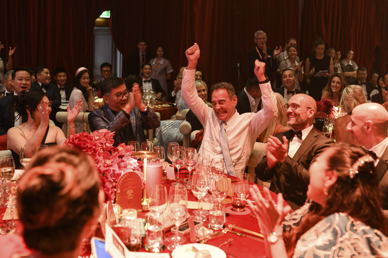 A man cheeering with his hands in the air seated at a dining table. The crowd of people around him are celebrating, cheering and clapping with him