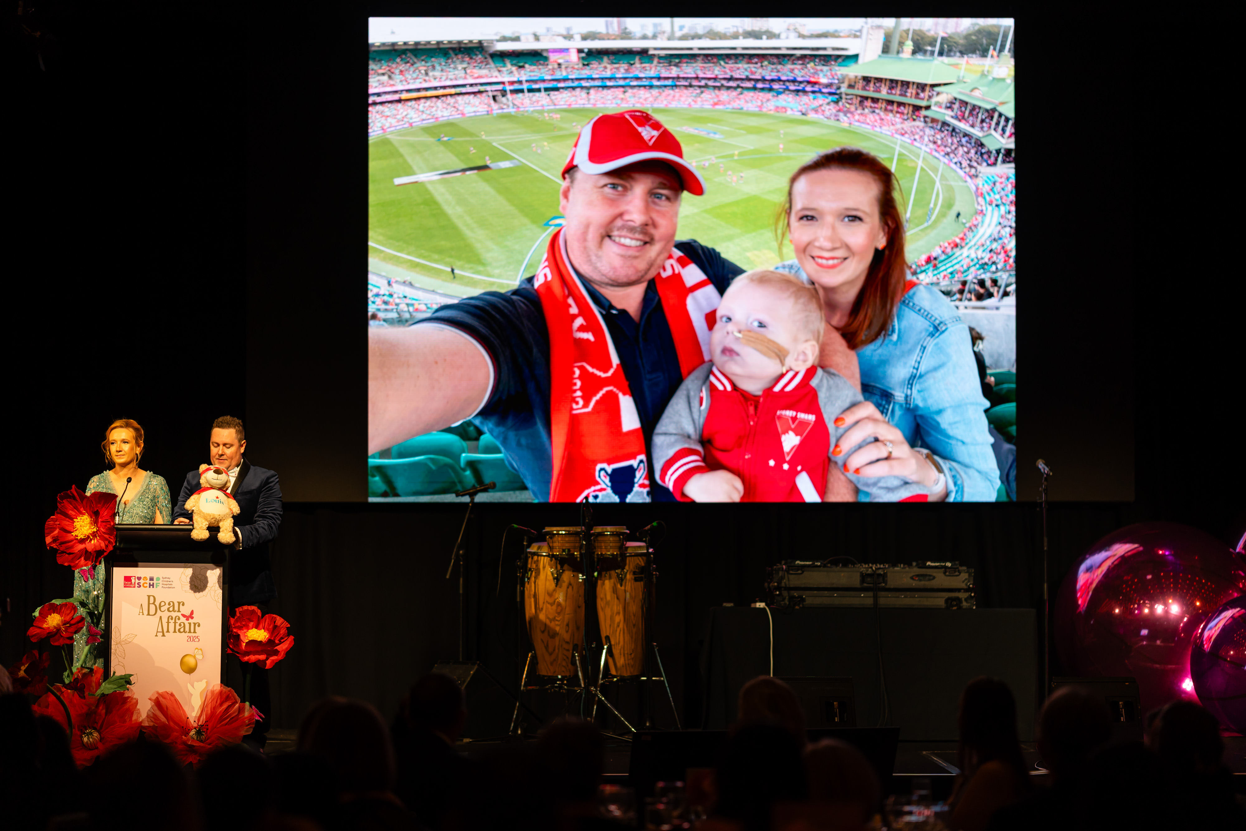 A family wearing red scarves taking a selfie in stadium seating, shown on a large event screen.