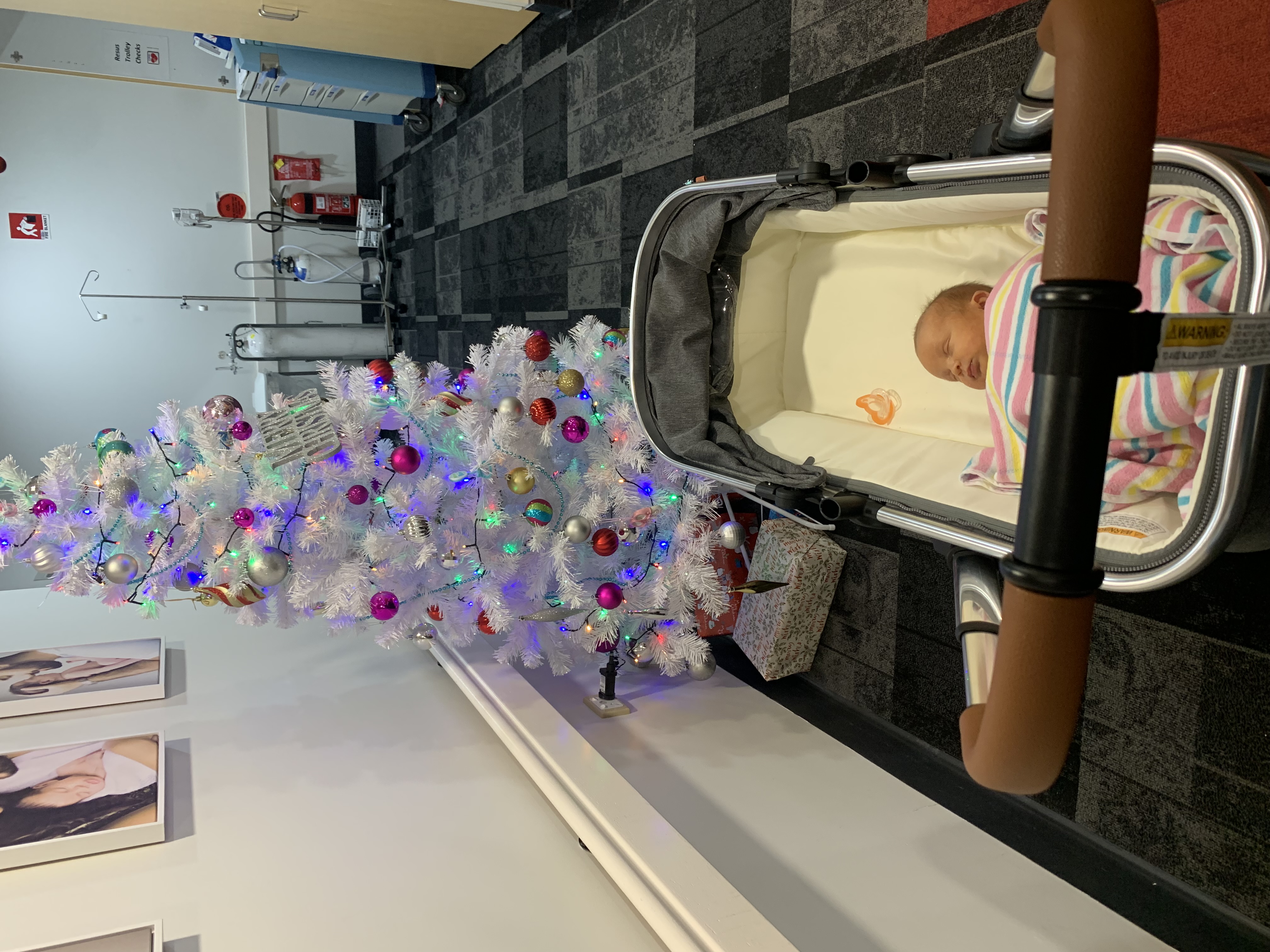 Image of baby in pram wrapped in hospital blanket, beside white Christmas tree.