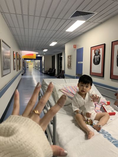 A child sitting on a hospital bed in a brightly lit hospital corridor, with a hand in the foreground reaching toward the child. Medical equipment, artwork on the walls, and hallway seating are visible in the background.