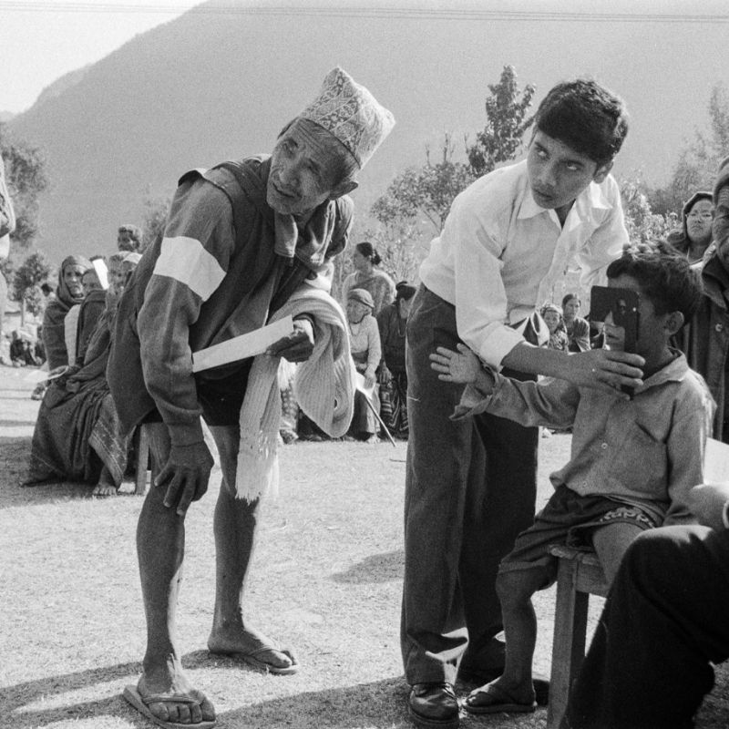 A black-and-white photograph of two men standing looking at something out of frame. One of the men is holding an eye-care device for a young boy who sits on a stool next to him, looking through the device.
