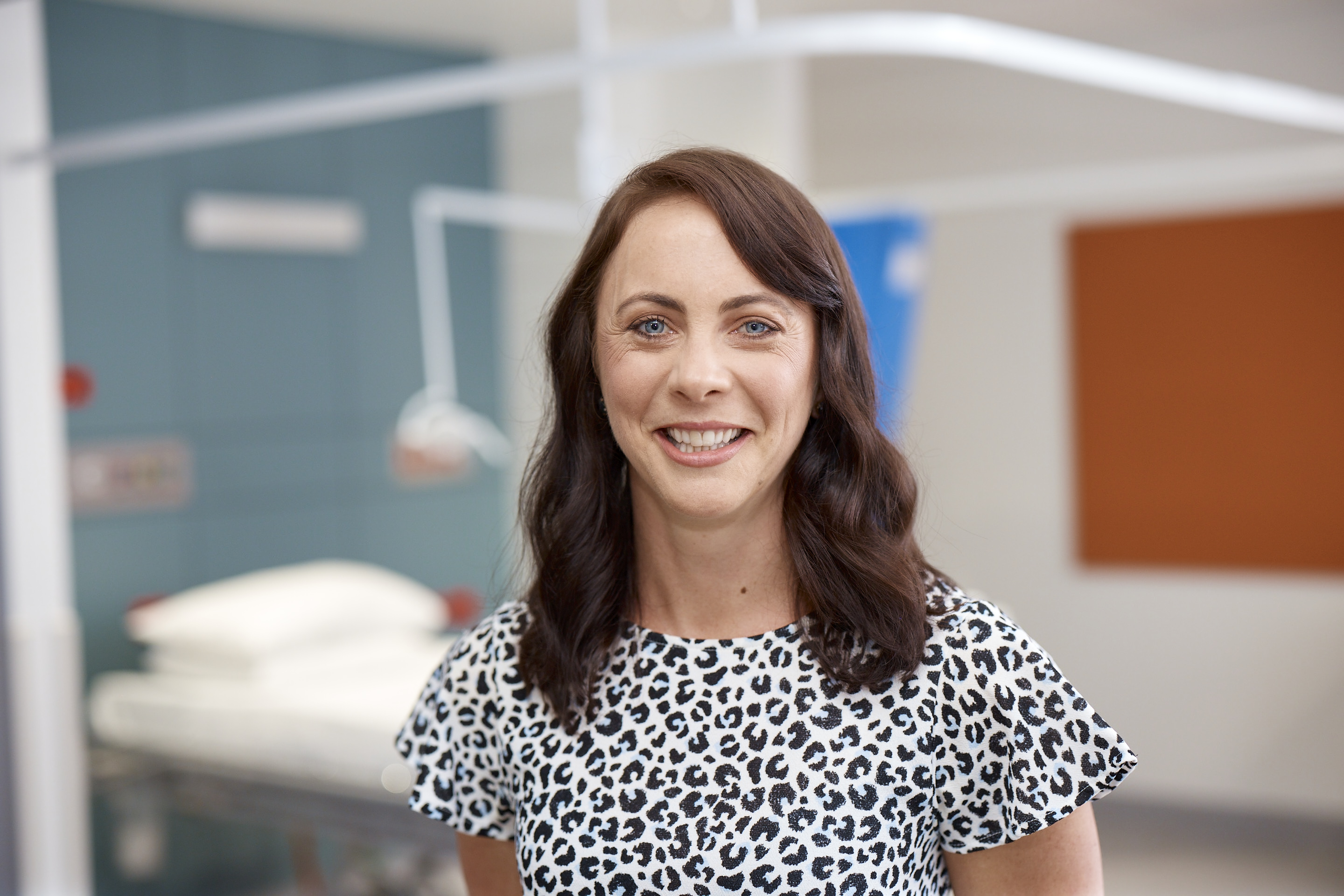 Person wearing a white and black leopard-print top standing in a hospital or clinical room with a bed and medical equipment in the background.