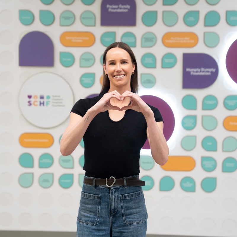 Emma Mckeon smiling while making a heart gesture with her hands, standing in front of a media wall branded with SCHF messaging and colours