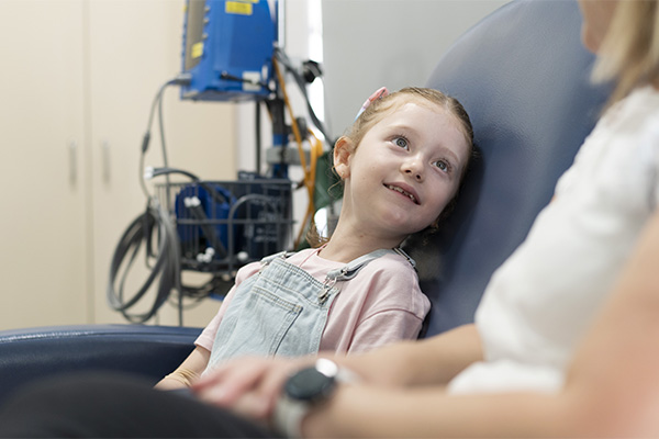 Bec sitting next to her mum in a hospital chair, with a mobile hospital equipment rack behind her