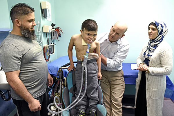 A boy with a breathing apparatus attached to his neck in a hospital room with his parents looking on as a doctor treats the boy.