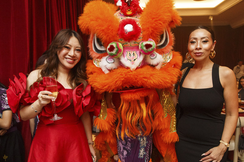 Two women in cocktail attire, one holding a glass of wine, with a person in a brightly coloured chinese dragon costume between them