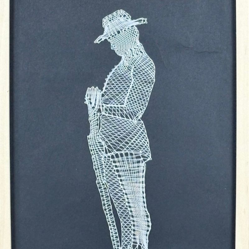 Photograph of a black canvas with an artwork of white lace intricately laid out in the shape of an ANZAC soldier's side silhouette. His head tilted down wearing a wide-brimmed hat as he holds his service weapon's hilt as the tip of the gun rests on the ground.