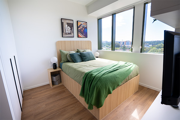 a bedroom with a wooden bedframe, bed with deep green sheets and pillows, two side tables with black and white rounded lamps and two print artworks hanging above the bedhead. Sydney's skyline can be seen out the three large windows next to the bed.