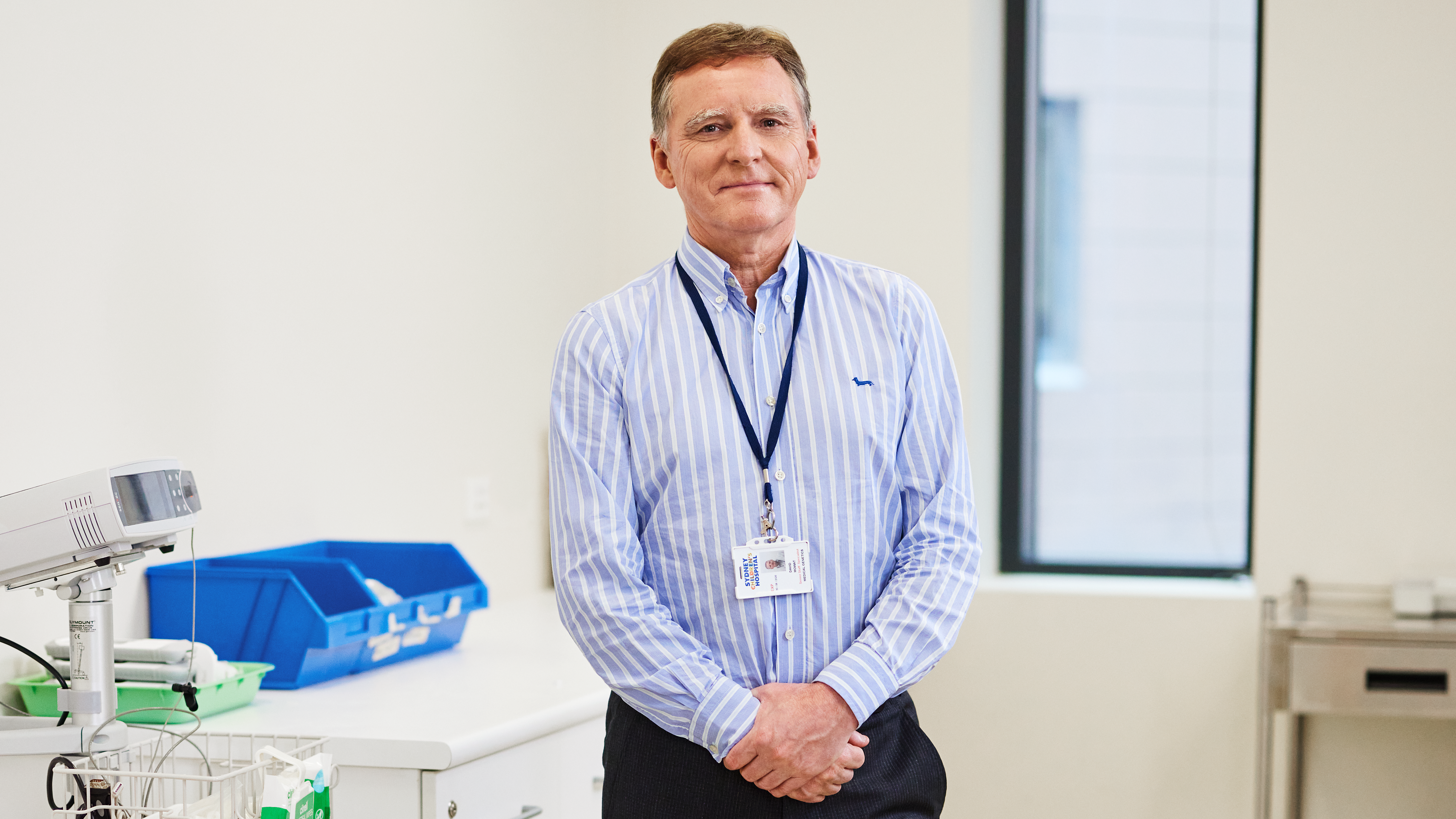 Person wearing a blue striped shirt and hospital ID badge standing in a clinical setting with medical equipment and storage trays in the background.