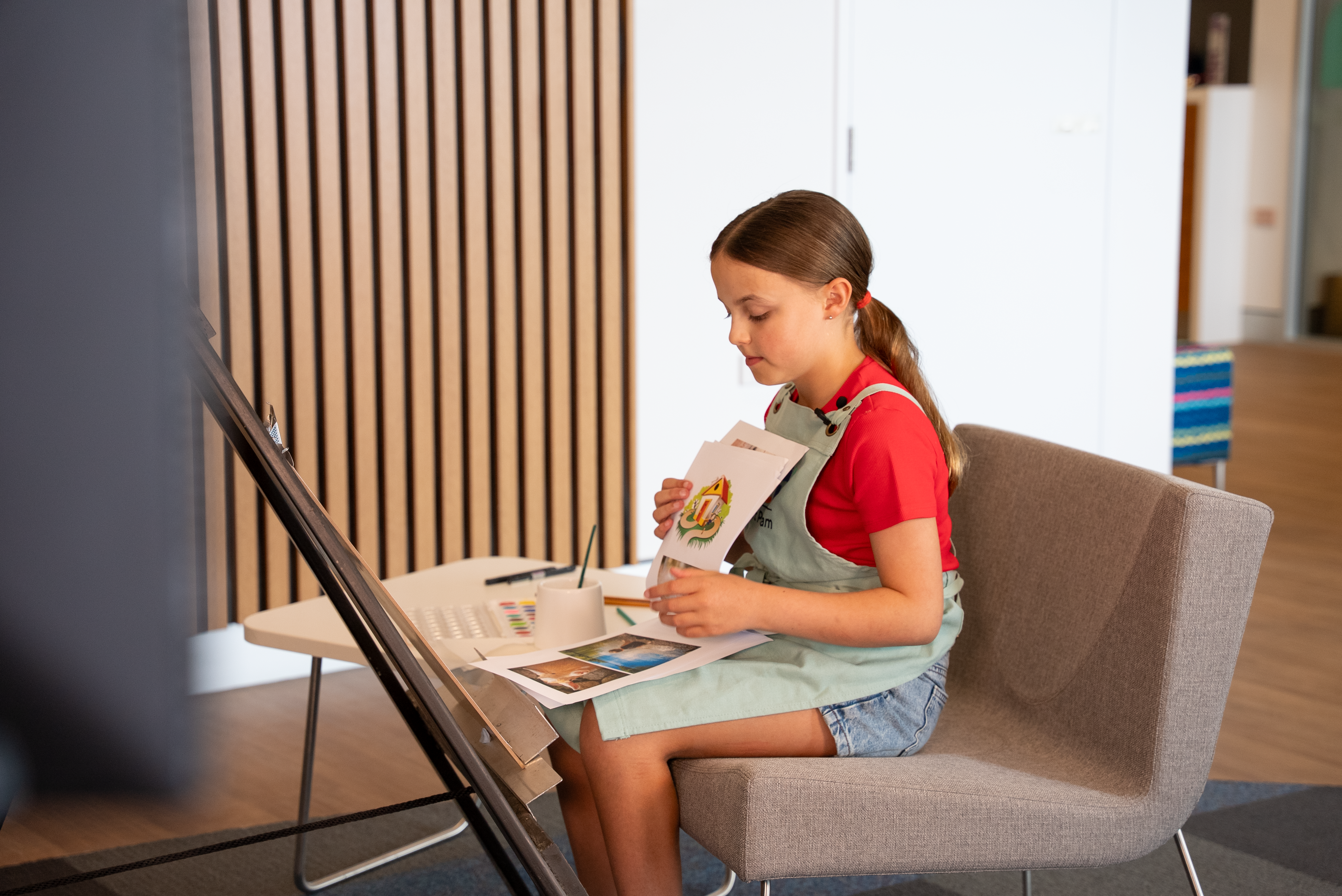 Child sitting on a chair wearing a red shirt and apron, holding a drawing while other art materials and papers are on a small table nearby.