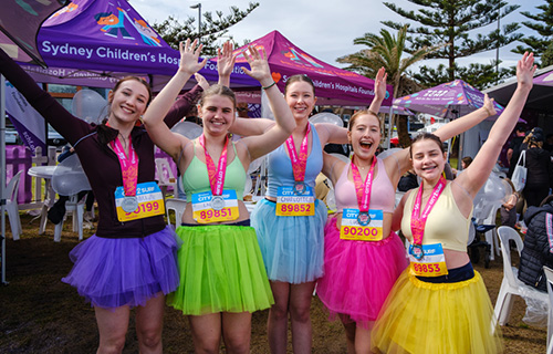 Five women wearing brightly coloured tutus sporting City2Surf medals around their necks all celebrating after the event