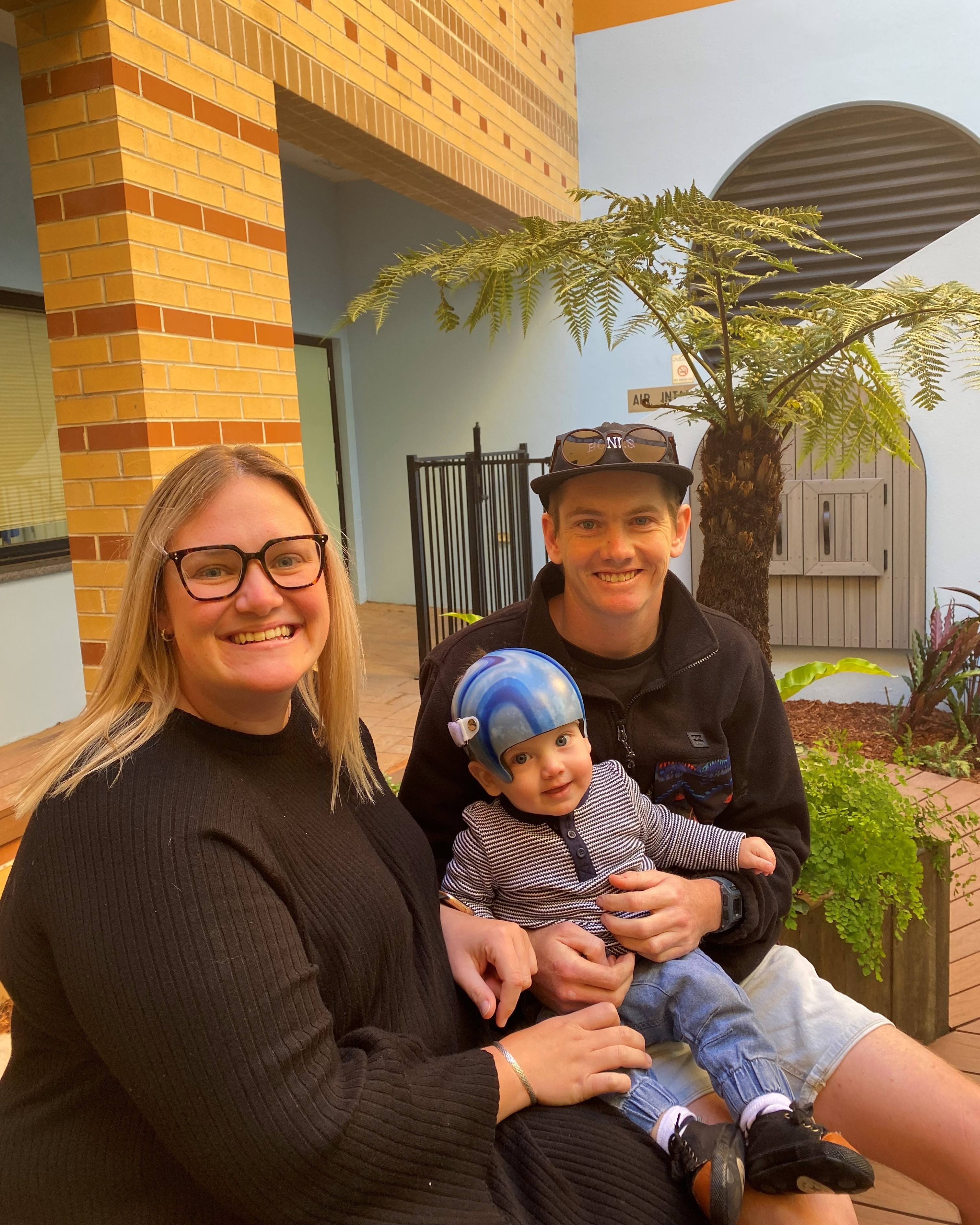 Three people sitting outside near a brick wall, with one holding a child wearing a blue helmet. Three people sitting outside near a brick wall, with one holding a child wearing a blue helmet.