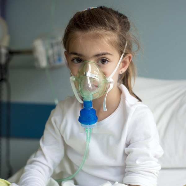 A young girl in a hospital bed wearing a breathing mask with a tube connected to some medical equipment behind her