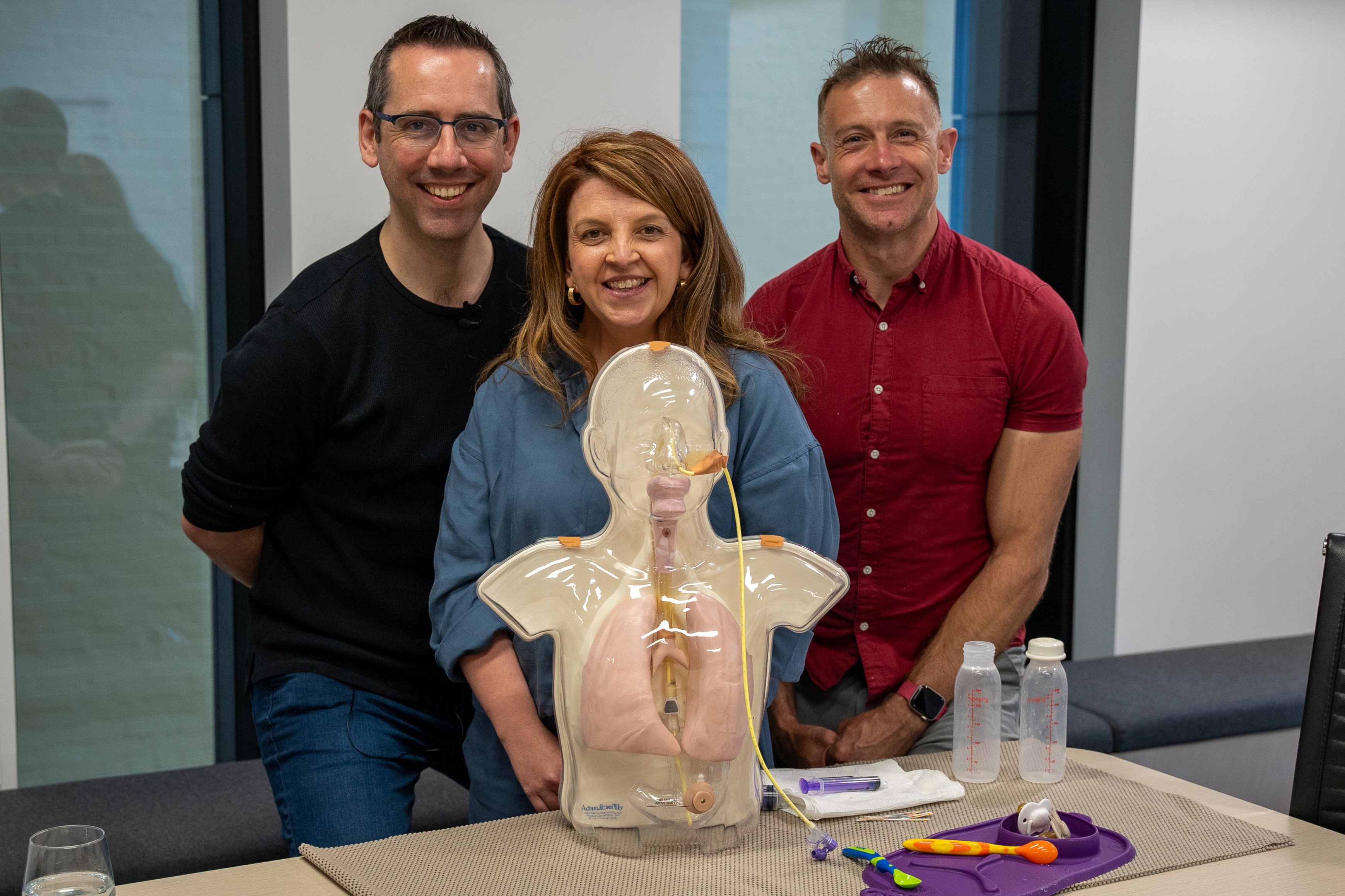 Three smiling adults stand behind a clear anatomical training mannequin of a child used for demonstrating tube feeding.