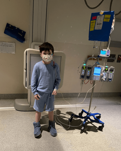 Cooper standing in a hospital hallway wearing his pajamas. Next to him is a trolley with lots of medical equipment and screens connected to wires monitoring him