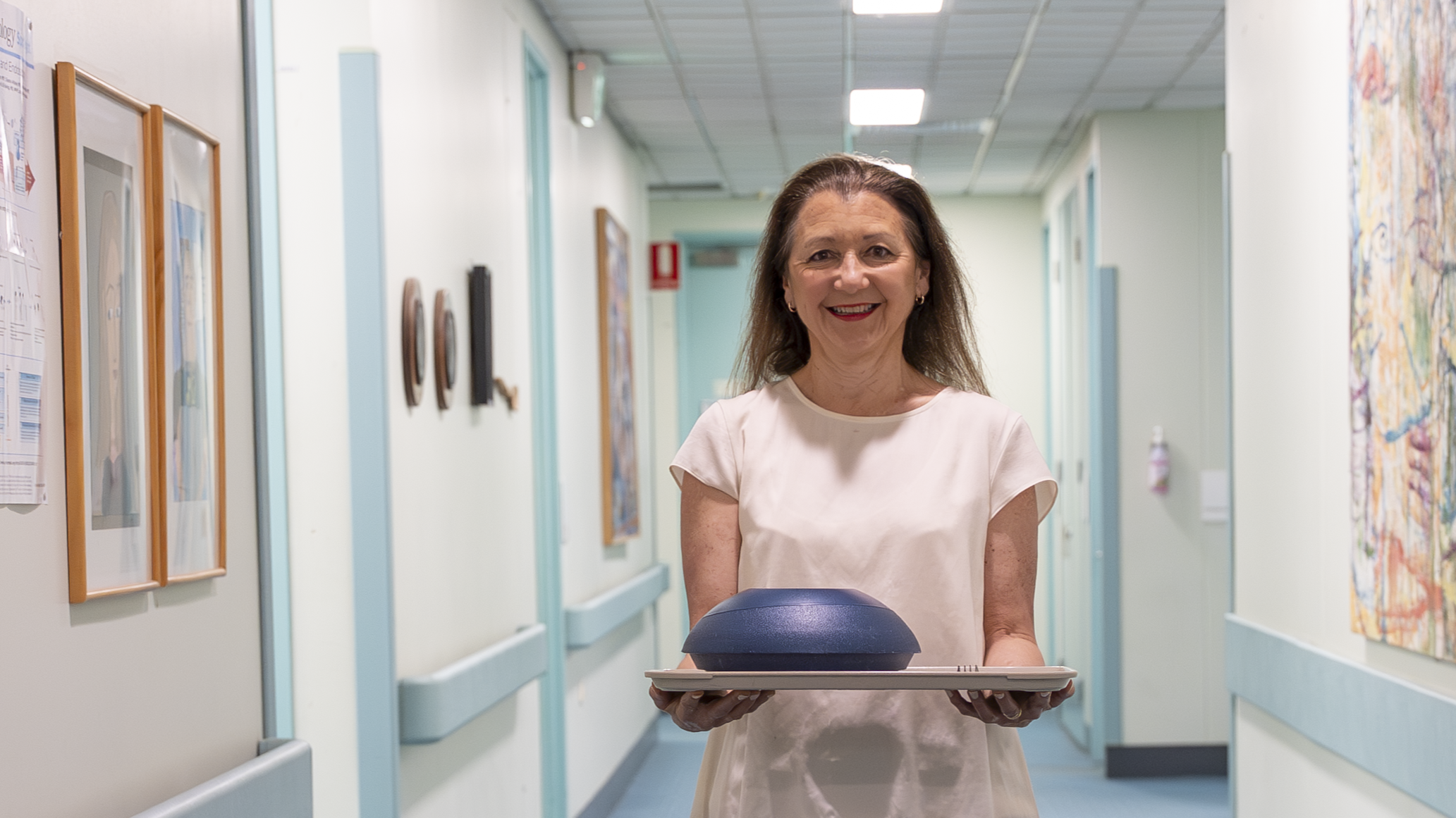 Person standing in a hospital corridor holding a tray with a blue bowl, surrounded by framed artwork and light-colored walls.