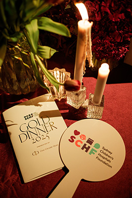 A table with tall, thin lit candles, an auction bidding paddle with an SCHF logo and a booklet with the words 'Gold Dinner 2025' written on it.