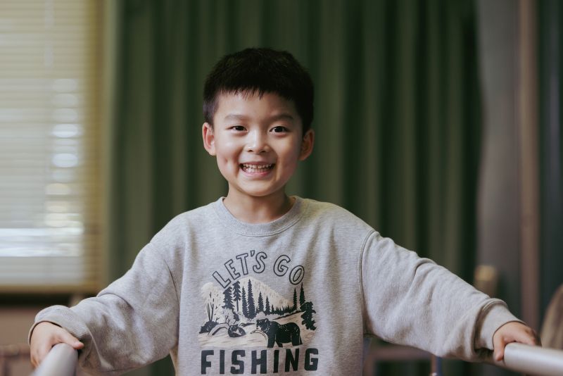 A child standing indoors holding onto parallel support bars, wearing a light grey “Let’s Go Fishing” sweatshirt with an illustrated outdoor scene.