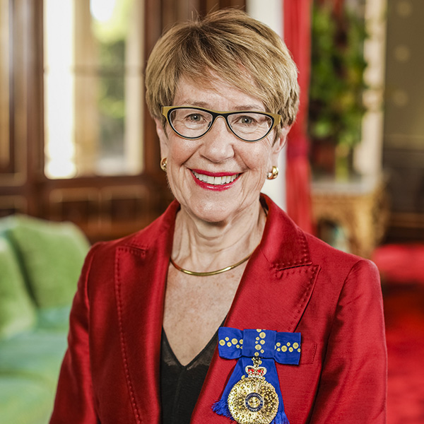 Her Excellency the Honourable Margaret Beazley AC KC standing in front of a bright green lounge and decorative timber furniture. She wears a red suit jacket and has a gold medal and blue ribbon pinned to her chest Her Excellency the Honourable Margaret Beazley AC KC standing in front of a bright green lounge and decorative timber furniture. She wears a red suit jacket and has a gold medal and blue ribbon pinned to her chest