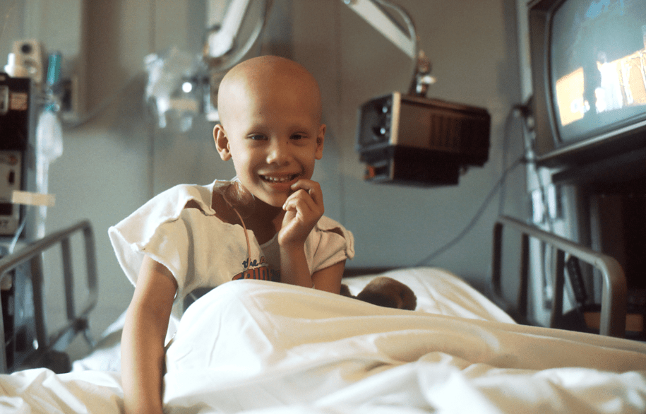 A children's hospital patient with a shaved head smiling while sitting in a hospital bed with white sheets. Behind them many pieces of medical equipment can be seen.