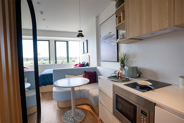 a kitchenette and attached breakfast nook with a curved lounge and small round table. Behind is a bedroom with lots of natural light flowing in through large wall length windows.