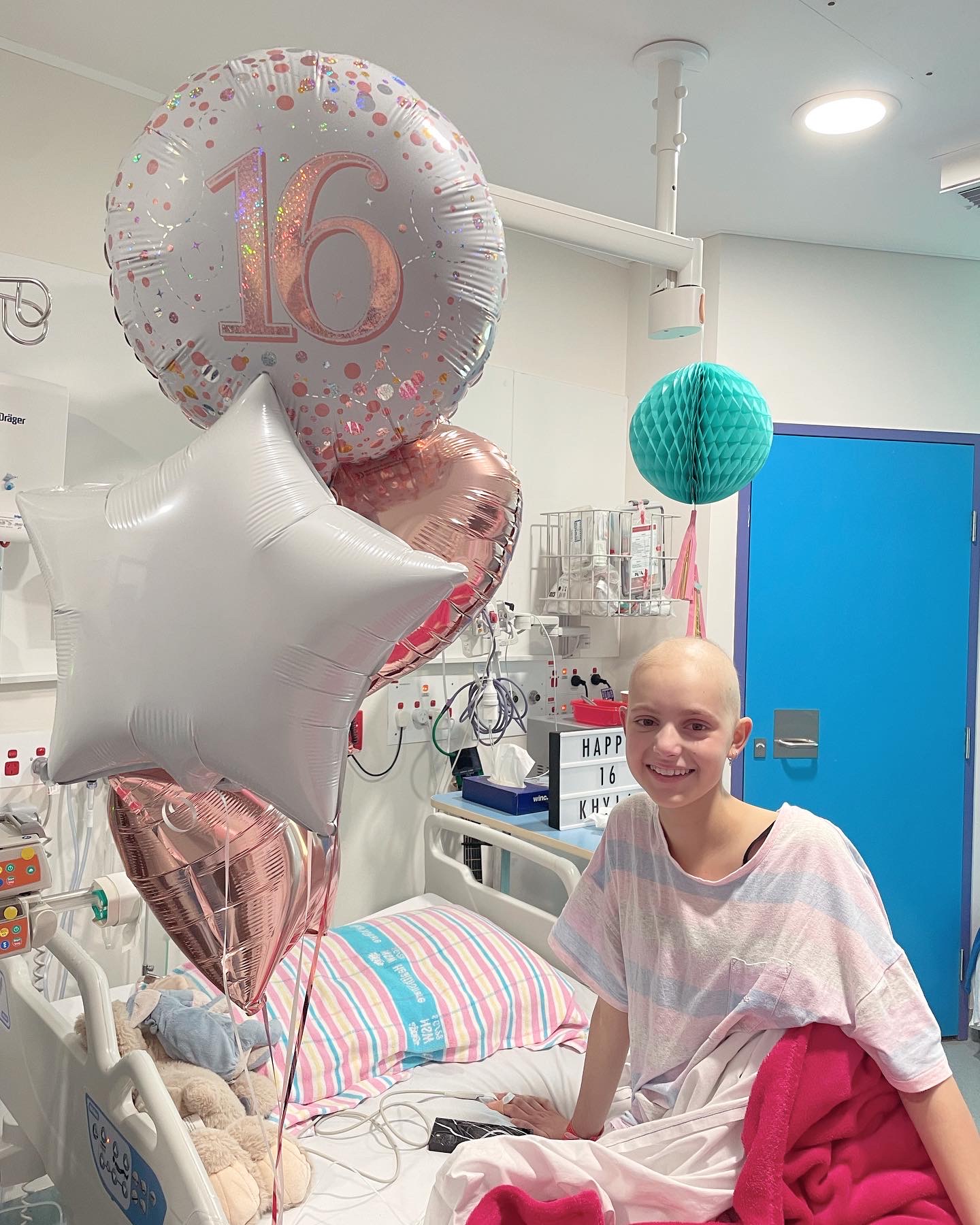 Kyla in a hospital bed with a bunch of balloons, one of which has a number 16 on it.