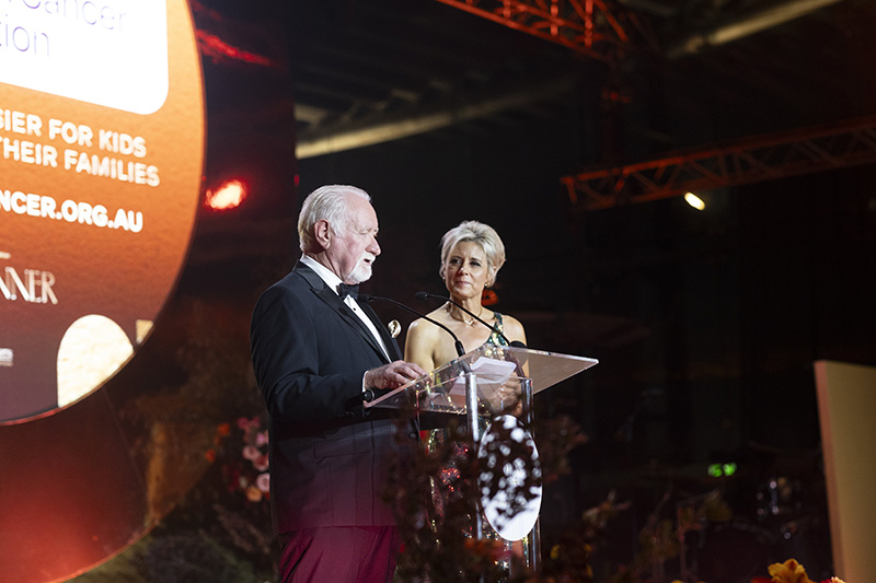 Well dressed man and woman standing at a podium together, speaking into microphones addressing an audience at a fancy gala dinner with bright stage lights shining on them and the golden backdrop behind them