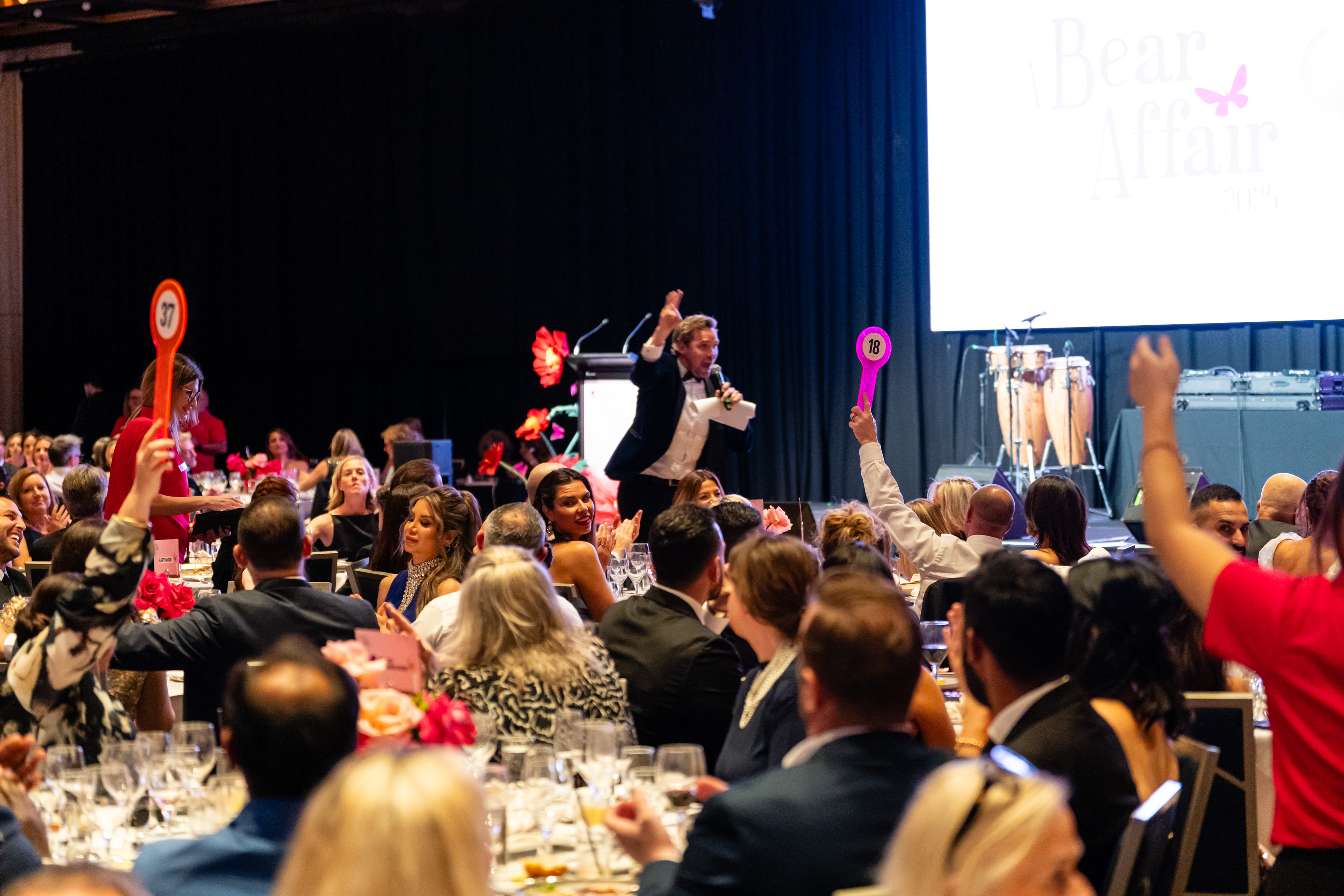 An auctioneer engaging the audience as guests raise bidding paddles at tables.