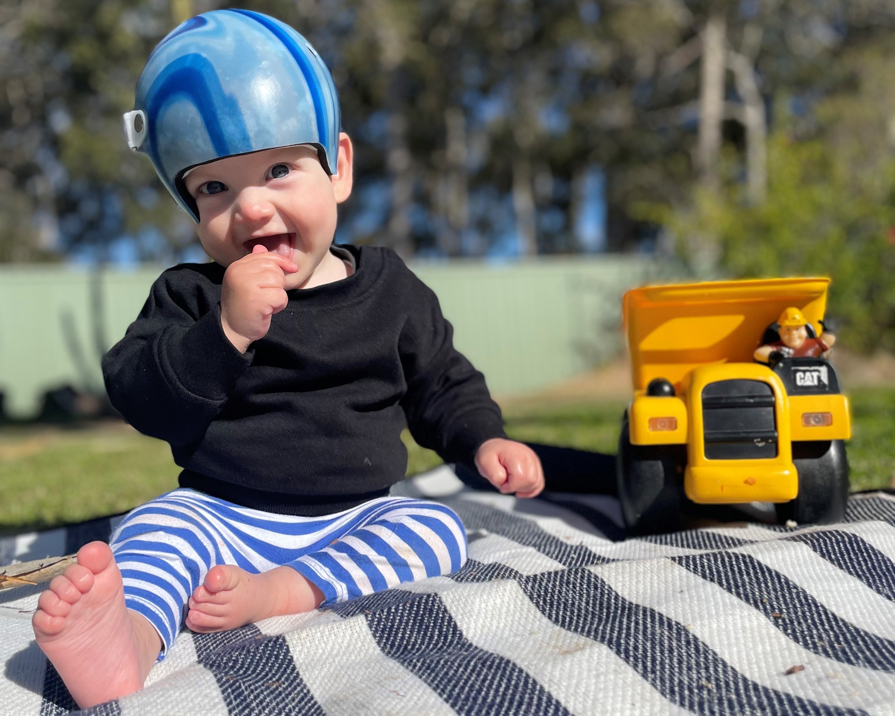 Smiling child in a helmet sits barefoot on a striped blanket outdoors beside a yellow toy truck.