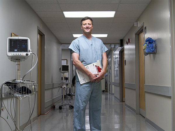 A male doctor holding a clipboard standing in the middle of a long hospital hallway with some medical monitoring equipment in the background