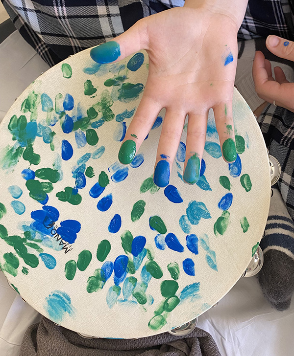 Close up of a child's hand with blue and green paint on their finger tips. Underneath resting on their lap is a tambourine that has many blue and green coloured paint dots all over it