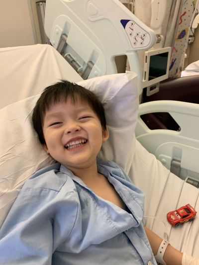 A child lying in a hospital bed wearing a light blue hospital gown, with medical equipment and another hospital bed in the background. A small toy car is placed beside the child on the bed.