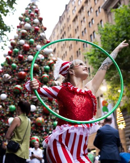 Image of festive dancer holding a hula hoop and dancing in front of large Christmas tree.