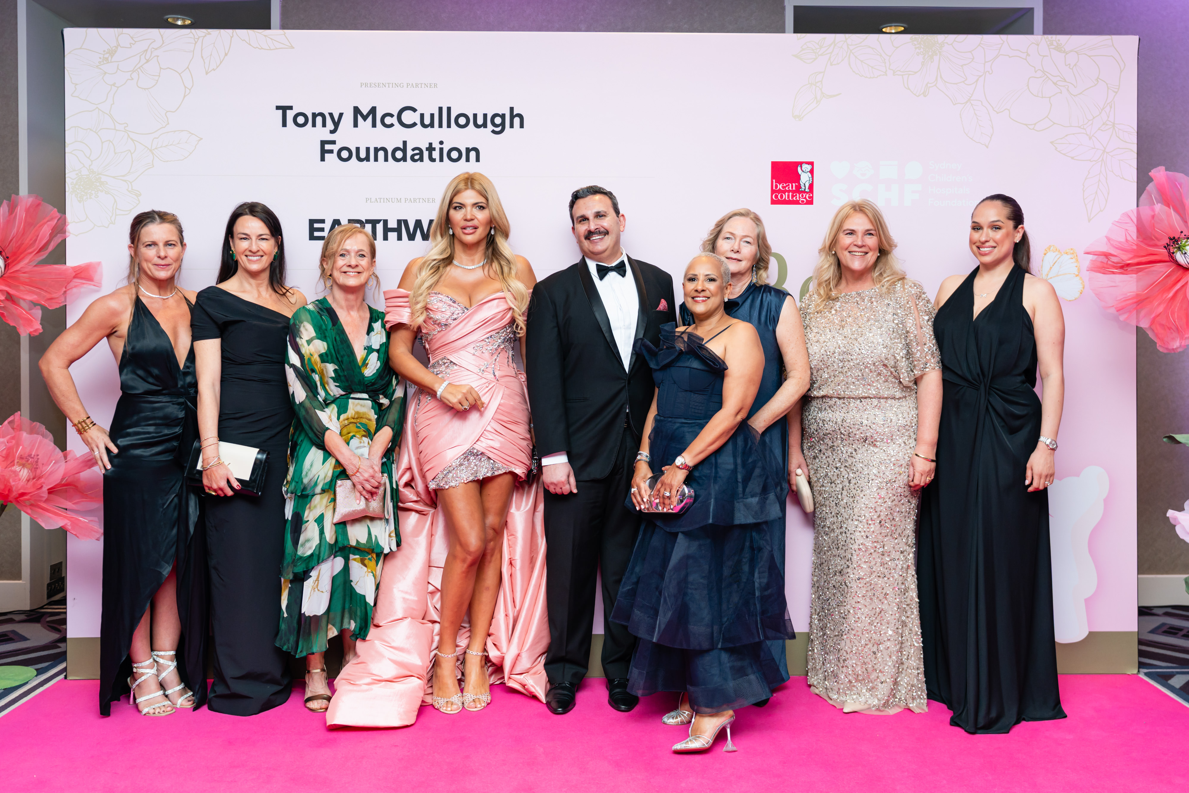 A group of formally dressed attendees standing together on a pink carpet in front of an event backdrop.