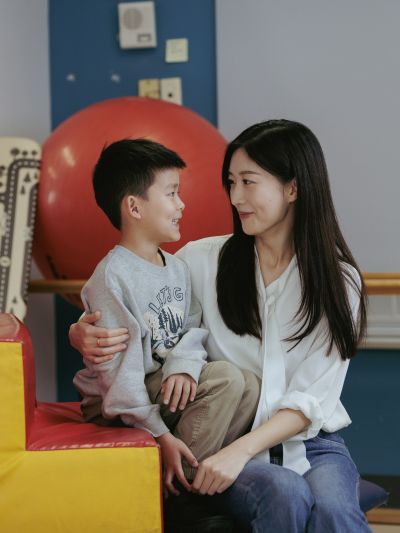 An adult seated beside a child on padded play equipment, with the adult’s arm around the child for support. A large red therapy ball and other playroom equipment are visible in the background.