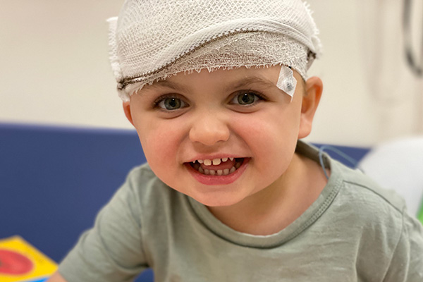 A young boy in hospital smiling. He has bandages wrapped around his head covering some electrode discs used to measure brain activity.