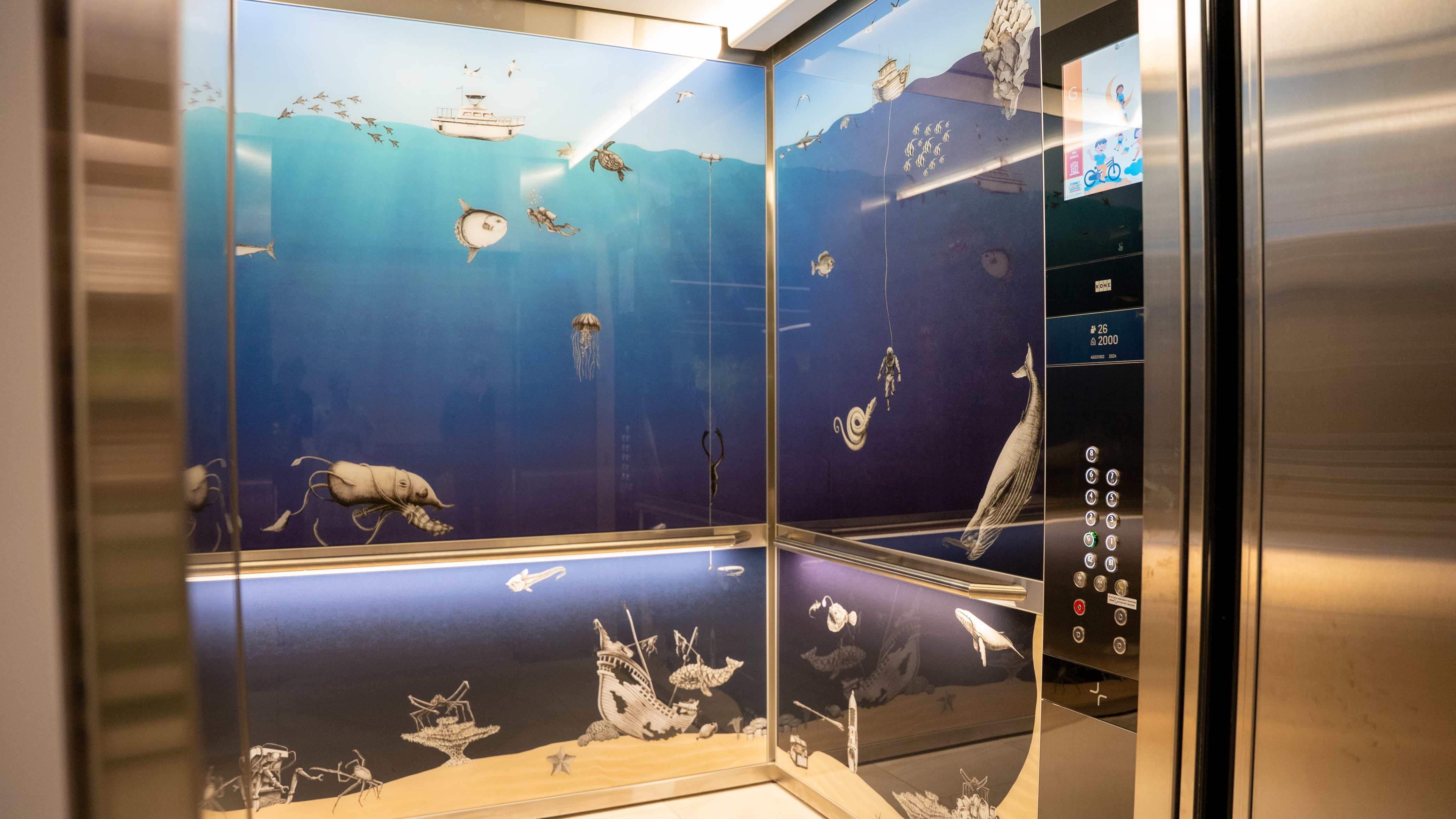 Elevator interior decorated with an underwater ocean theme, featuring illustrations of marine life such as fish, jellyfish, and whales, along with a sandy seabed and a boat on the water’s surface.