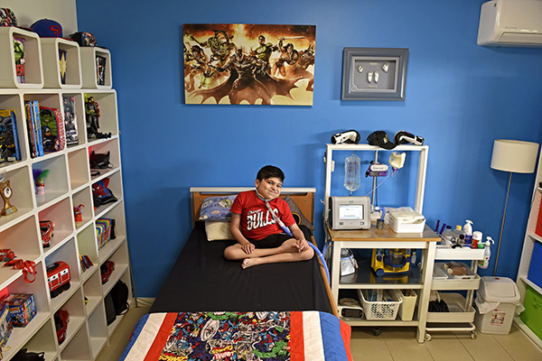 A boy with a neck-breathing apparatus sitting on his bed wearing a red t-shirt. His room has a mix of superhero toys and posters juxtaposed with medical equipment next to his bed