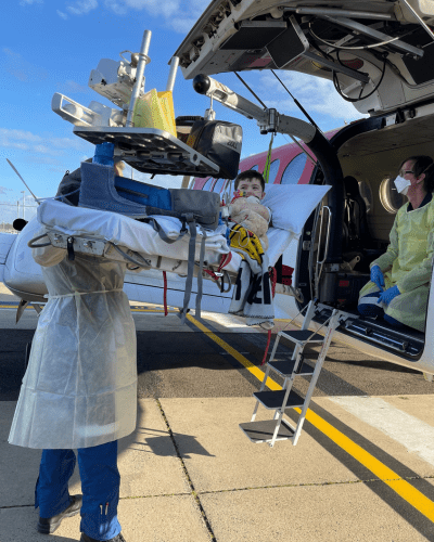 Cooper laying on a bed connected to a long metal arm that hoists him into the back of an aeroplane as he's transferred via plane to a different hospital