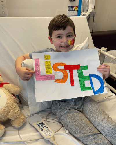 Cooper in his hospital bed holding up an artwork he made that says 'LISTED'