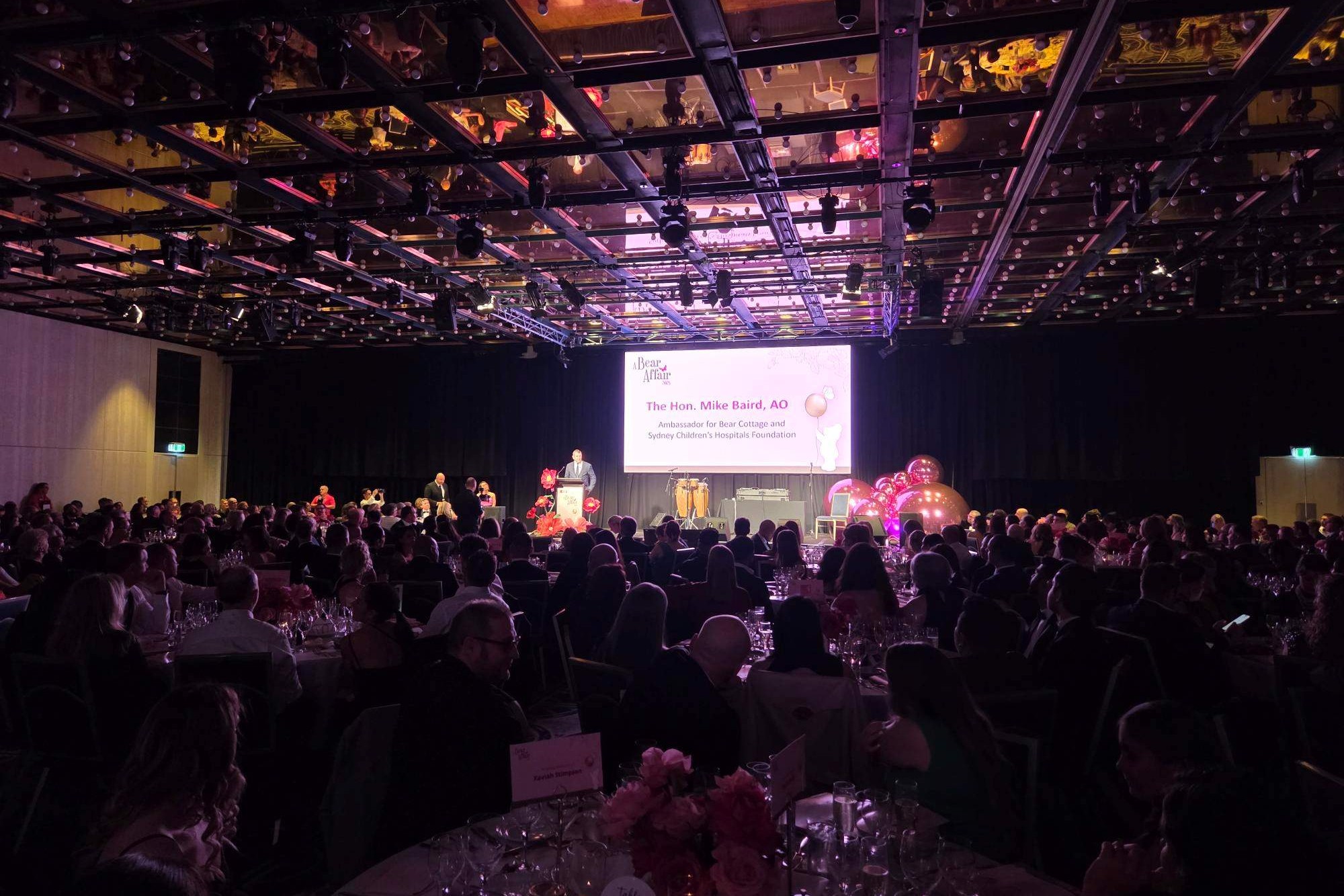 A large banquet room filled with seated guests facing a stage and presentation screen.