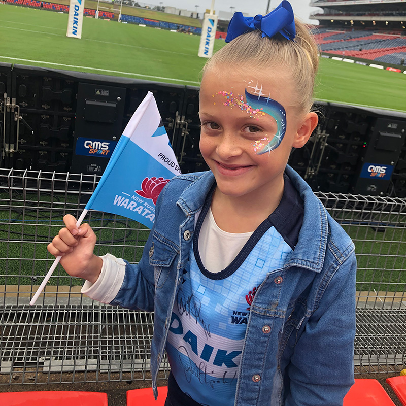 Olivia at a stadium wearing a Warratahs uniform and holding a Warratahs flag with facepaint on