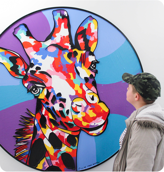 A boy wearing a camouflage hat looking at an artwork of a multi-coloured giraffe