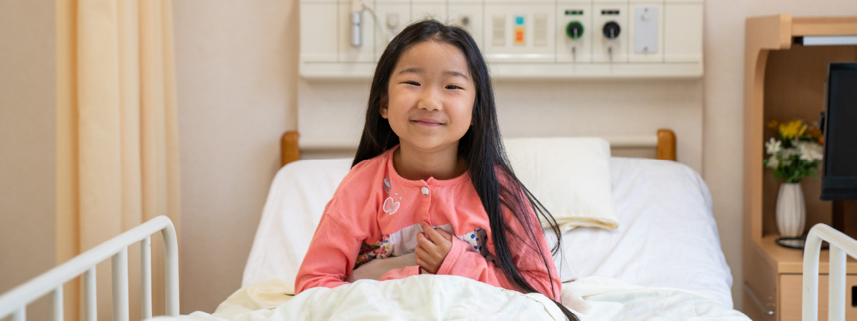A young girl sitting on a hospital bed smiling. She is wearing pink pajamas and looks happy.