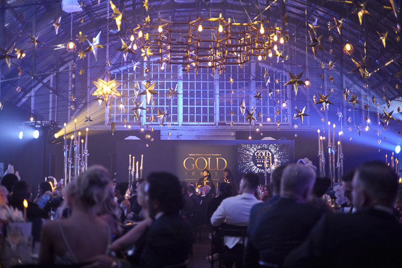 All guests seated at their tables with the stage in the background. Gold coloured ceiling decorations hang and shine in the bright lights. All guests seated at their tables with the stage in the background. Gold coloured ceiling decorations hang and shine in the bright lights.