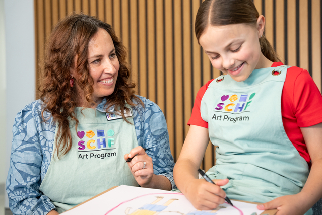 A woman and a young girl wearing SCHF Art Program aprons while they draw colourful artwork on cardboard backed paper