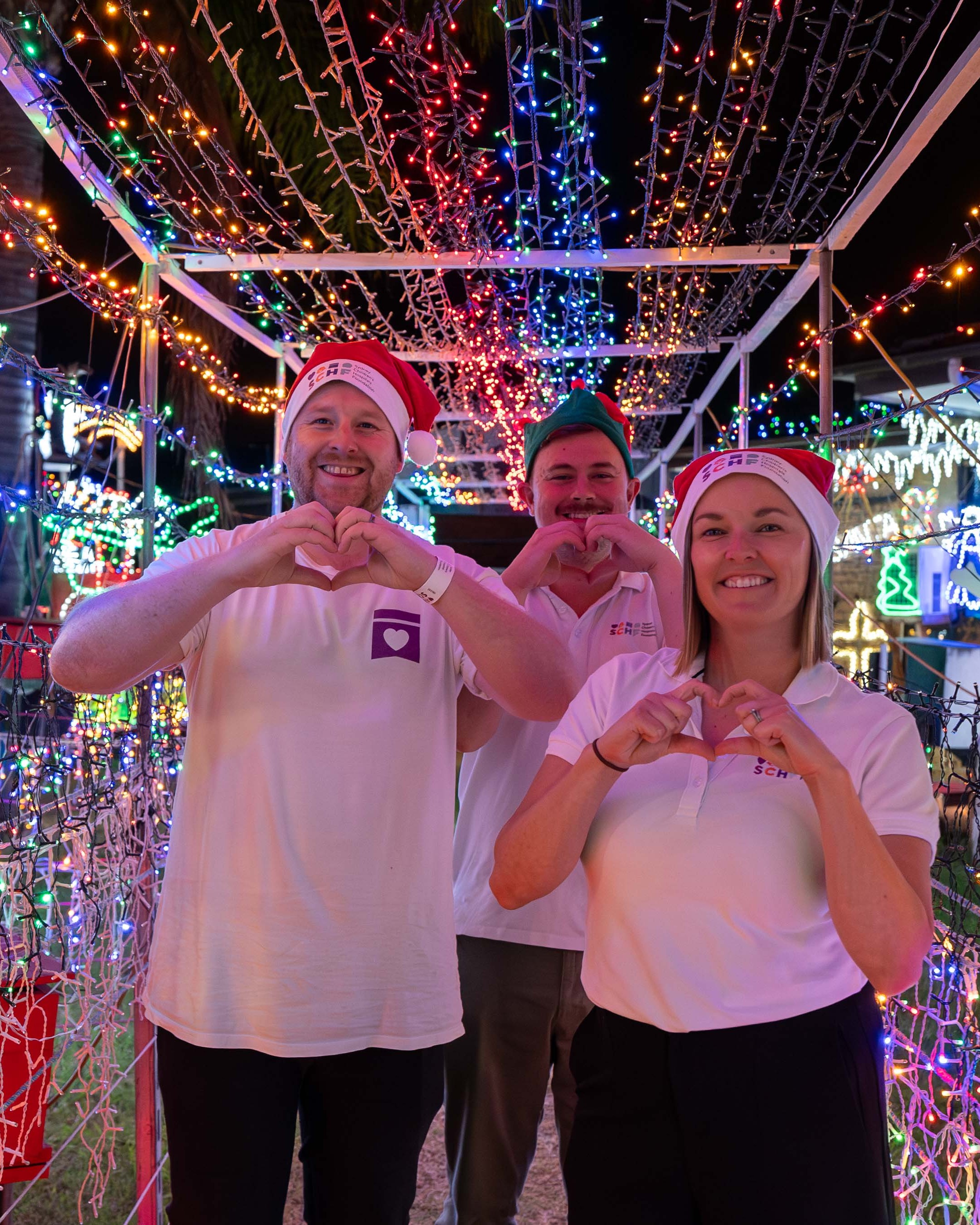 Image of three adults standing in Christmas light arch and doing heart shaped hands.