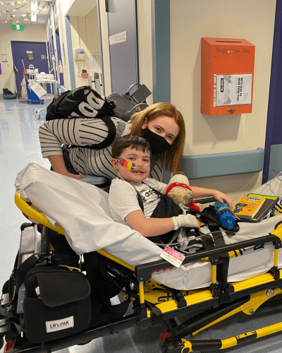 Cooper laying in a hospital stretcher with his mum behind him comforting him as he's moved through the hospital. Despite his situation, he is smiling broadly.